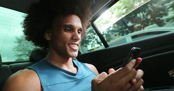 Black man in backseat of car typing on cellphone smiling, riding taxi while on smartphone