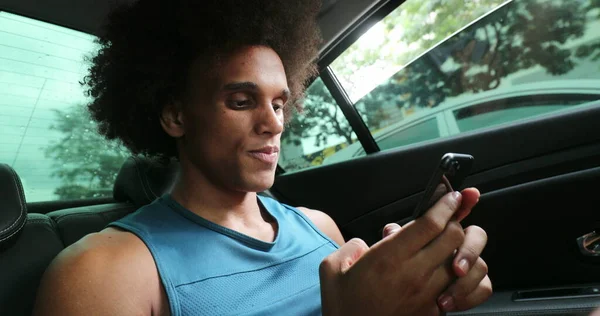 Black man in backseat of car typing on cellphone smiling, riding taxi while on smartphone