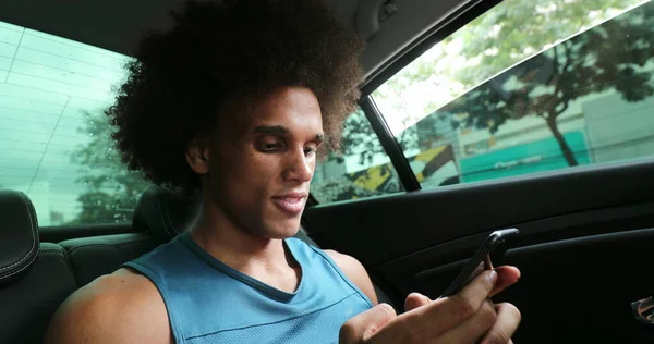 Black man in backseat of car typing on cellphone smiling, riding taxi while on smartphone