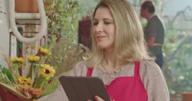 Female business owner of retail store using tablet technology to check inventory while employees work in the background. A middle aged business woman entrepreneur owner of small business store