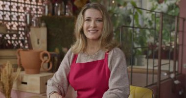 Flower shop business owner smiling at camera inside shop wearing apron. A middle aged female entrepreneur owner of local business