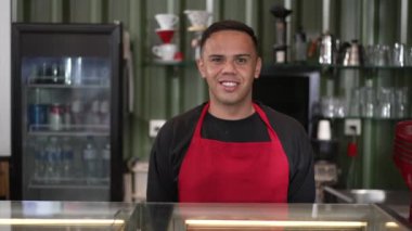 Kafenin arkasındaki Latin kahveci genç bir adamın portresi. Güney Amerikalı iş adamı kameraya gülümseyen kırmızı önlük giyiyor.