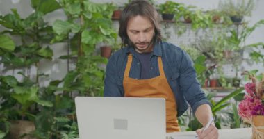 Young florist entrepreneur checking online orders smiling in front of laptop. Small business online orders concept. Male employee wearing apron writing down online orders