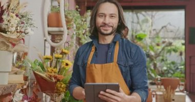 Portrait of a male employee holding tablet and wearing apron working at small business flower store. Young staff looking at camera smiling using modern digital technology