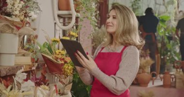 One happy middle aged female entrepreneur holding tablet standing inside flower shop checking for online orders with digital modern device. Small business owner checking inventory managing small business