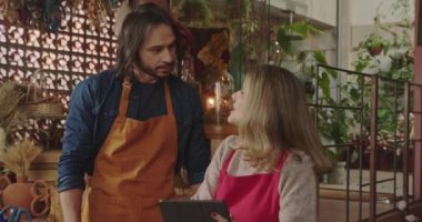 Local business owner of flower store talking with employee. Female entrepreneur managing shop talks with male employee wearing apron