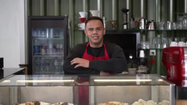Barista kameraya gülümsüyor. Kahve tezgahının arkasında kırmızı önlük giyen İspanyol bir çalışanın portresi. Küçük yerel işletme
