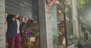 Businesswoman owner of small business store at sidewalk level opening storefront sliding shutters up. Open for business concept. Female entrepreneur opened for customers and clients