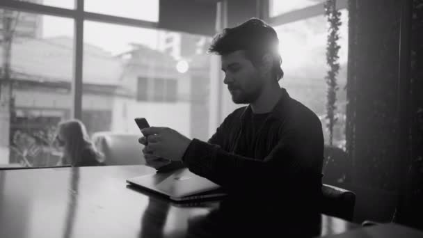 カフェでインターネットを使って携帯電話を見ている喫茶店の若い男 モノクロショット — ストック動画