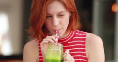 İki çok kültürlü kız yan yana oturmuş yeşil detoks sıvısı içiyor ve cep telefonu cihazlarını kontrol ediyor, gülüyor ve gülümsüyor.