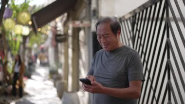 Happy senior man using cellphone standing outdoors in city street. Portrait older Asian American person typing message on smartphone device smiling