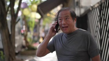 Happy senior man receiving great news on phone standing in city street. Authentic real life laugh and smile of an older Asian person speaking with cellphone laughing