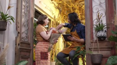 Two work colleagues of small local business checking online orders inventory. Young woman holding beautiful bouquet flower arrangement showing to worker holding tablet