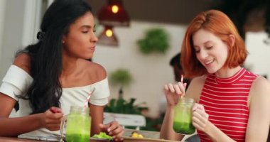 Çekici iki genç çok ırklı kadın yemek yiyor ve taze yeşil smoothie içiyor.