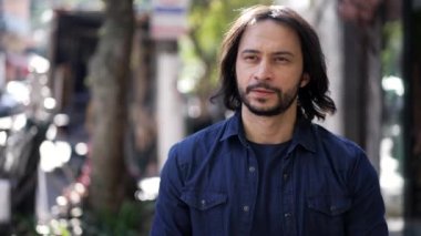 Pensive young casual man walking in street outdoors. Thoughtful person in 30s walks in urban environment. Male with long hair
