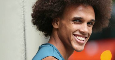 Happy mixed race handsome young man smiling to camera while leaning on wall in city setting
