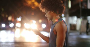 Young african man walking in city street at night looking at cellphone device