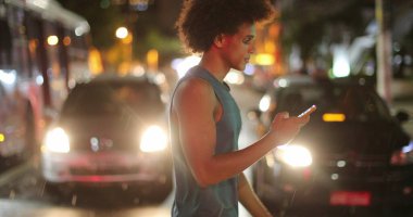 Young african man walking at night looking at cellphone device while crossing street during rain