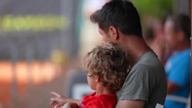 Baba ve çocuk birlikte, küçük çocuk babasının kucağında oturup tenis maçı izliyor.