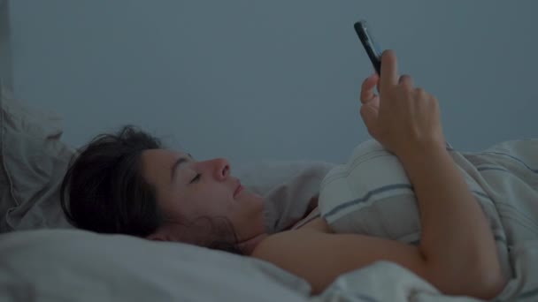 女性はベッドで寝そべって朝のベッドでスマートフォンを見て カジュアルと現実の生活 — ストック動画
