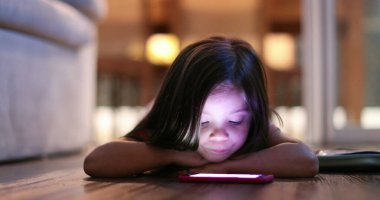 Child watching cellphone screen at night. Blue light from smartphone device glowing on little girl face