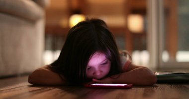 Little girl staring at cellphone screen at night. Child watching smartphone lying on floor at home