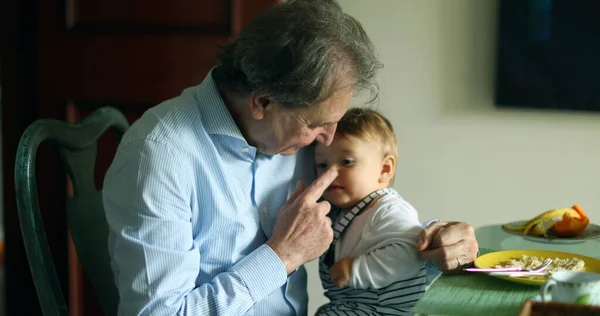 Grandfather holding grandson in arms at meal table. Casual and candid grand-parent with infant baby