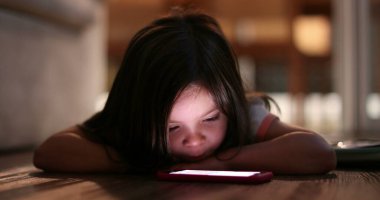 Small girl staring at cellphone screen at night. Cute kid lying on floor watching cellphone screen