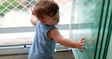 Baby standing by apartment window during lockdown, infant child exploring home window