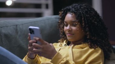 Evde oturma odasındaki kanepede akıllı telefon kullanan rahat bir Afrikalı Amerikalı kız. Geceleri telefona bakan siyahi bir genç kadının yakın çekimi.