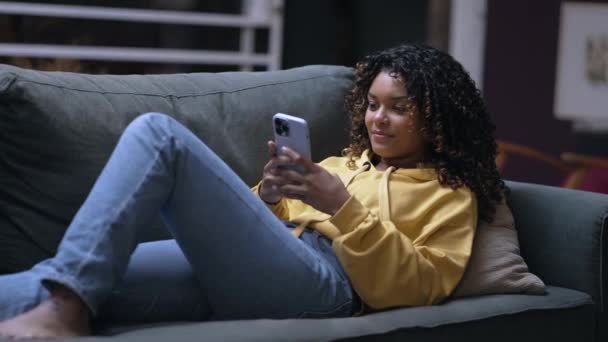 ある若い黒人女性がソファに座ってスマートフォンを見ていた 携帯電話を使ったリラックスしたアフリカ系アメリカ人の20代の少女 ソーシャルメディアをチェックする者 — ストック動画