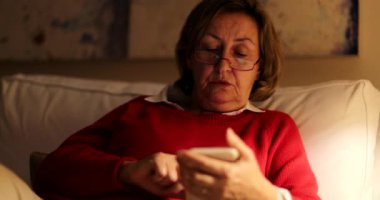 Akşamları akıllı telefon ekranına bakan yaşlı insan evdeki kanepede oturuyor.