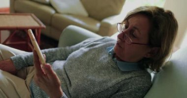 Kanepede oturan akıllı telefon cihazından içeriği izleyen kıdemli bir kadın.