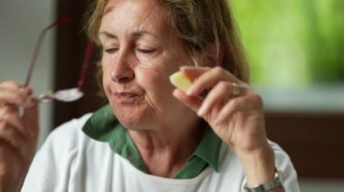 Düşünceli, yaşlı bir kadın yemek çiğnerken okuma gözlüğünü çıkartıyor ve derin derin düşünüyor.