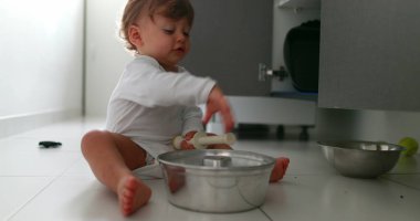 Baby playing with pans and pots on kitchen floor. One year old toddler infant hitting metal utensil