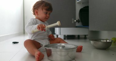 Baby playing with pans and pots on kitchen floor. One year old toddler infant hitting metal utensil