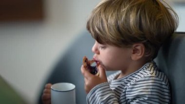 Toddler eating bread with jelly and drinking from mug in breakfast