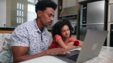 Ev mutfak masasında dizüstü bilgisayarın önünde siyah bir baba ve kız.