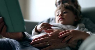 Kanepede otururken tablet kullanırken kucağında bebek tutan büyük anne.