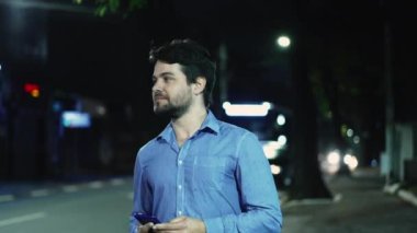 Kaygısız genç adam, yağmurlu bir yağmurda gecenin bir yarısı elinde telefonla şehir kaldırımında yürüyor. Mutlu adam dışarıda yürüyor.