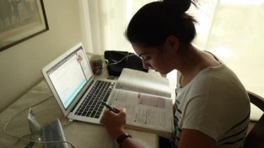 Sınav için evde okuyan genç bir kadın. Masasında açık bilgisayar ve kitap olan biri.