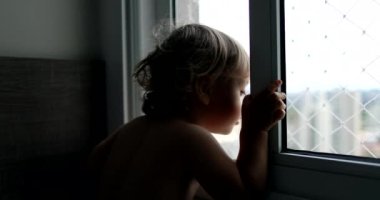 Küçük çocuk apartmanın penceresinin yanında durmuş dışarıya bakıyor.