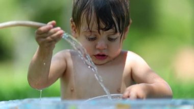 Sevimli bebek arka bahçedeki plastik havuzda su hortumuyla oynuyor.