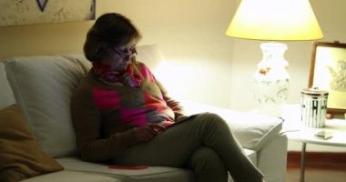 Altmışlı yaşlarında, elinde tabletle kanepeye oturmuş interneti seyreden samimi bir kadın.