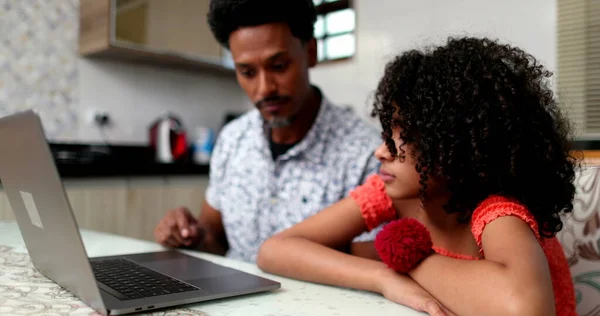 Black father helping daughter with computer laptop at home kitchen