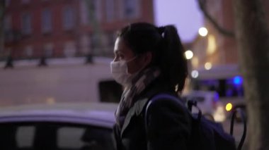 30 'lu yaşlarda bir kadın gece kaldırımda yürürken maske takıyor.