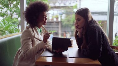 Kahve dükkanında çalışan iki farklı genç kadın birlikte tablet ekrana bakıyor. Siyahi bir kız, kadın meslektaşına işi anlatıyor.