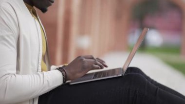 Dışarıda oturan siyah bir adam bilgisayarda yazı yazıyor. Afrikalı kişi uzaktan çalışma