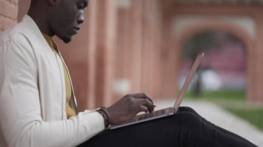 Dışarıda oturan siyah bir adam bilgisayarda yazı yazıyor. Afrikalı kişi uzaktan çalışma