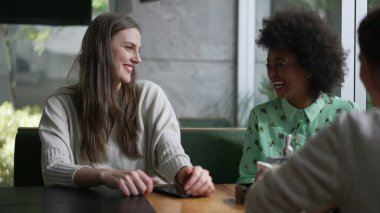 Kahve masasında oturmuş gülen ve gülümseyen üç farklı genç kadın. Konuşurken kız arkadaşlar arasındaki etkileşim. Gerçek hayatta gül ve gülümse.